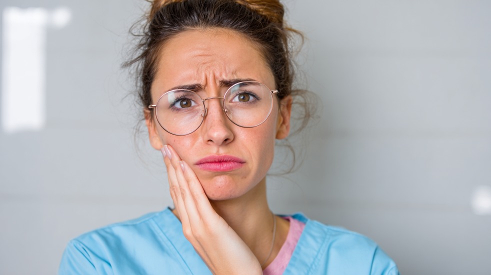 En kvinne i blå uniform og briller holder hånden mot kinnet og ser ut til å ha sterke tann- eller kjevesmerter