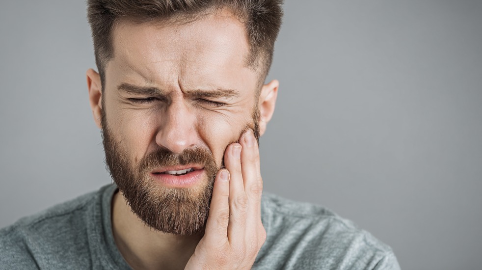 En mann med skjegg og grå t-skjorte holder hånden mot kinnet og ser ut til å oppleve smerte eller nummenhet i tannen
