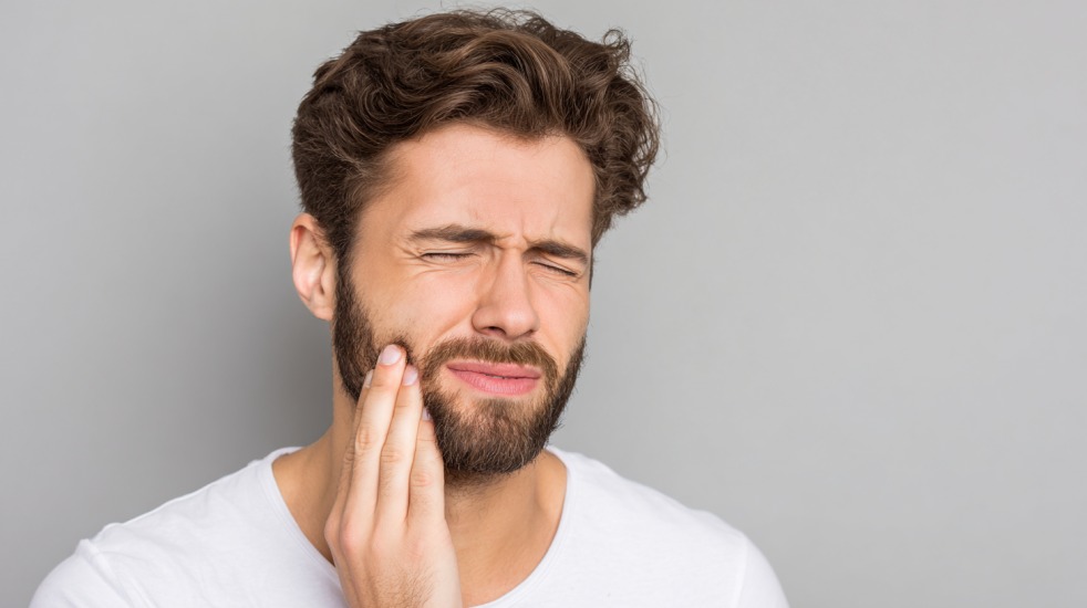 En mann med skjegg og hvit t-skjorte holder hånden mot kinnet og ser ut til å ha tann- eller kjevesmerter